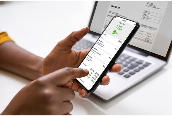 Person, die ein Smartphone hält und eine bezahlte Rechnung darauf anzeigt. Auf dem Bildschirm des Smartphones ist eine Rechnung mit einem grünen Häkchen und dem Text "$325 Paid" sichtbar. Im Hintergrund ist ein Laptop mit einem offenen Dokument zu sehen, das ebenfalls eine Rechnung darstellt. Die Hand der Person zeigt mit dem Finger auf eine spezifische Stelle auf dem Smartphone-Bildschirm, wodurch die Interaktion mit der digitalen Rechnung hervorgehoben wird.